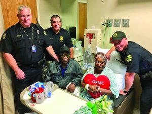 Three smiling officers surround an older woman in a hospital bed. 